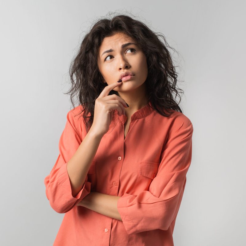 pretty young woman thinking about problem, confused emotion, isolated, wearing orange shirt, hipster style, curly hair, holding finger at lips, looking up, having idea